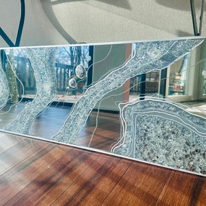 May include: Rectangular mirror with a silver frame. The mirror's surface features a design of layered, textured, and sparkling silver and gray elements, outlined with white lines. The mirror is displayed on a wooden surface.