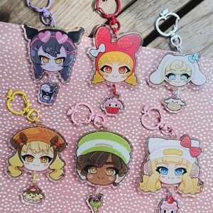 Op de afbeelding: Zes acryl sleutelhangers met gestileerde anime-karakterhoofden en bijpassende appelvormige sluitingen. Elk karakter heeft een kleine bungelende bedel die bij het ontwerp past, waaronder een drankje, cupcake, gebak en dessertitems.