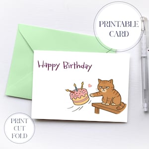 Może przedstawiać: Drukowana kartka urodzinowa z kreskówkowym kotem popychającym ciasto ze stołu. Na karcie widnieje napis "Happy Birthday" w fioletowym skrypcie. Na zdjęciu widnieje również jasnozielona koperta i długopis.