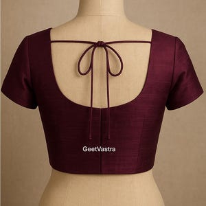 May include: A burgundy blouse with short sleeves, a scoop back, and a tie closure. The garment is displayed on a mannequin. The text "GeetVastra" is visible on the lower center of the blouse.