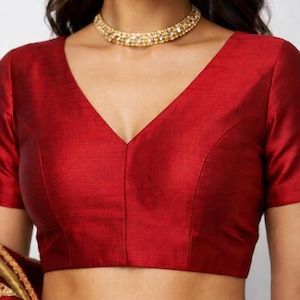 May include: A red, short-sleeved blouse with a deep V-neck. The blouse has a smooth, satin-like texture and is a solid, rich red color. A gold necklace is visible at the neckline. The blouse is part of a traditional garment.