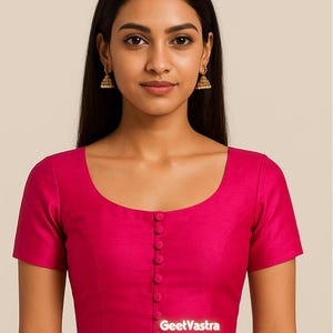 May include: A magenta blouse with short sleeves and a scoop neckline. The blouse has a row of buttons down the front and the text "GeetVastra" at the bottom. The model is wearing gold earrings.