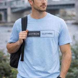 Könnte beinhalten: Hellblaues T-Shirt mit kurzen Ärmeln und dem Text "AVYRON CLOTHING" in einem schwarz-weißen Rechteck-Design. Das T-Shirt ist aus weichem Material. Die Person trägt einen schwarzen Rucksack.
