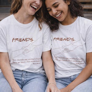 Könnte beinhalten: Zwei Personen tragen weiße T-Shirts mit dem Wort "FRIENDS" und einer Grafik von zwei Händen, die sich halten. Die Shirts haben auch ein Zitat über Freundschaft. Sie tragen blaue Jeans.