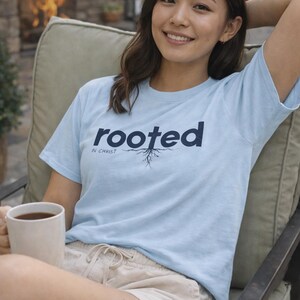 Könnte beinhalten: Hellblaues T-Shirt mit dem Wort "rooted" in Marineblau und dem Satz "in Christ" darunter. Eine Grafik von Wurzeln befindet sich unter dem Wort "rooted". Die Person sitzt in einem Stuhl und hält eine weiße Tasse.