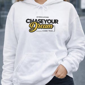 Könnte beinhalten: Weißer Kapuzenpullover mit Fronttasche und Kordelzug. Der Hoodie zeigt den Text "CHASE YOUR DREAM - COME TRUE." in schwarzer und gelber Schrift. Der Markenname "AVYRON CLOTHING" steht darüber.