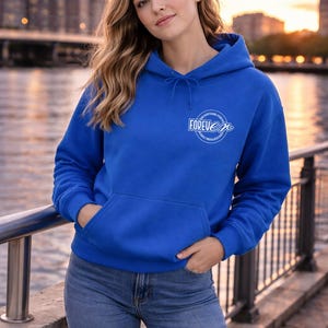 Könnte beinhalten: Ein königsblauer Kapuzenpullover mit einer Vordertasche und einer Kapuze mit Kordelzug. Der Kapuzenpullover hat das Wort "FOREVER" in einem weißen Kreisdesign auf der Brust. Die Person trägt eine blaue Jeans. Der Hintergrund zeigt eine Skyline bei Sonnenuntergang.