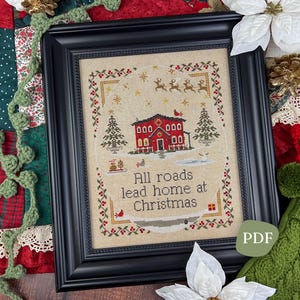 May include: Framed cross-stitch art with a black frame. The artwork features a red house, Christmas trees, and the text "All roads lead home at Christmas." The design includes reindeer and snowflakes, evoking a festive holiday theme.