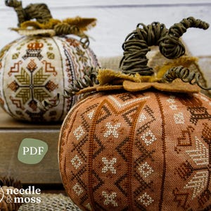 Puede incluir: Dos calabazas de tela con intrincado bordado de punto de cruz en tonos marrón, naranja y crema. Cada calabaza tiene un tallo de alambre verde retorcido y un detalle de hoja de fieltro. Un icono verde con el texto PDF y un logo con el texto needle & moss.
