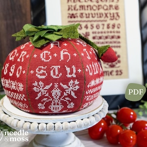 Op de afbeelding: Een rode, ronde, kruissteekversiering met witte letters en sneeuwvlokontwerpen. De versiering heeft een groene stoffen steel en staat op een witte, met kralen versierde taartstandaard. Een ingelijste sampler staat op de achtergrond.
