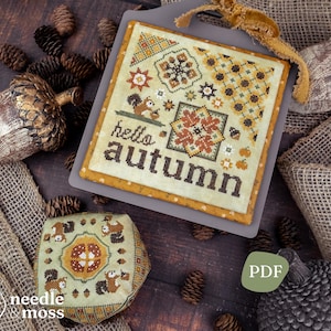 May include: A square cross-stitch sampler featuring the text hello autumn in dark brown letters, surrounded by motifs of a squirrel, acorns, pumpkins, and geometric floral patterns in shades of orange, brown, green, and yellow on cream fabric.