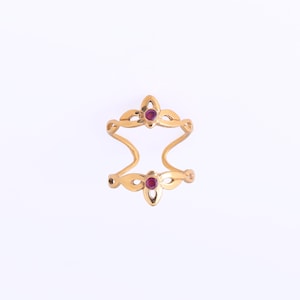 Könnte beinhalten: Ein goldfarbener Doppel-Fingerring mit floralem Design. Der Ring hat zwei rubinrote Edelsteine, einen oben und einen unten, mit einem offenen Design dazwischen. Der Ring ist vor einem weißen Hintergrund platziert.