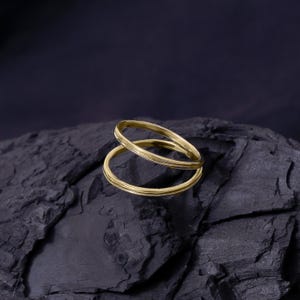 May include: Two gold-colored rings, one slightly overlapping the other, are displayed against a dark, textured background. The rings have a simple, elegant design, with a smooth, polished finish. The rings are likely jewelry.