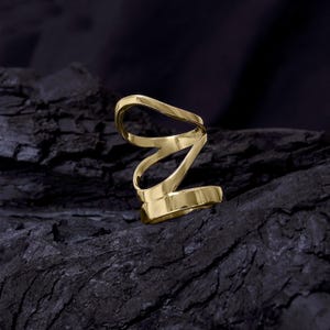 May include: A gold-colored, abstract ring with a swirling, open design. The ring is made of a shiny, reflective metal and sits on a dark, textured surface. The ring's design features a continuous, flowing shape.