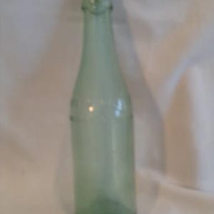May include: A vintage, pale green glass bottle with a long neck and rounded base. The bottle has a slightly textured surface and appears to be empty. The bottle is likely from the early to mid-20th century.