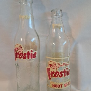May include: Two clear glass Frostie root beer bottles. The bottles have a red and white label with the word "Frostie" and a cartoon character. One bottle is taller than the other.