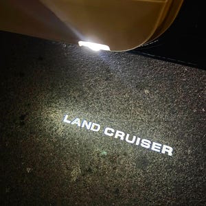 May include: Illuminated "LAND CRUISER" text on a dark, textured surface, likely a car's floor mat. A bright overhead light shines down from the car's interior, highlighting the text. The car door is visible in the background.