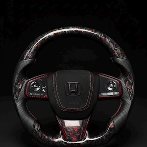 Puede incluir: Un volante Honda personalizado con un dise&ntilde;o de fibra de carbono negro y rojo. El volante presenta un agarre de cuero perforado, costuras rojas y el logotipo de Honda en el centro. El fondo es negro.