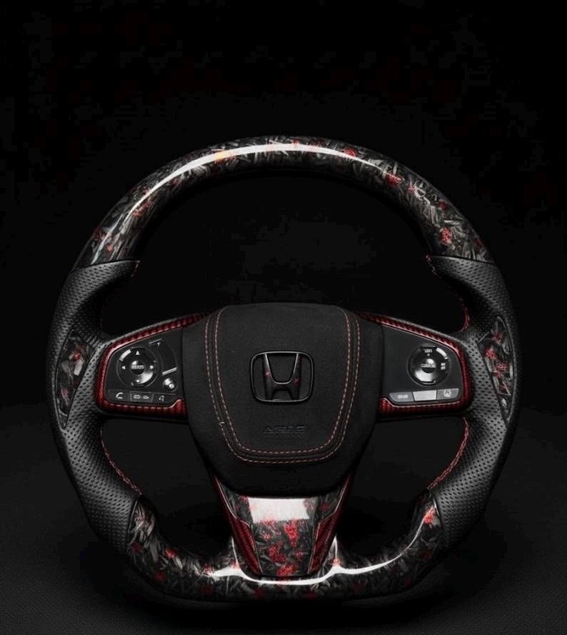 Puede incluir: Un volante Honda personalizado con un dise&ntilde;o de fibra de carbono negro y rojo. El volante presenta un agarre de cuero perforado, costuras rojas y el logotipo de Honda en el centro. El fondo es negro.