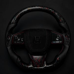 Puede incluir: Un volante Honda negro y rojo con un dise&ntilde;o de fibra de carbono. El volante presenta un agarre de cuero perforado con costuras rojas y controles integrados. El centro del volante tiene un acabado de gamuza negra con el logotipo de Honda.