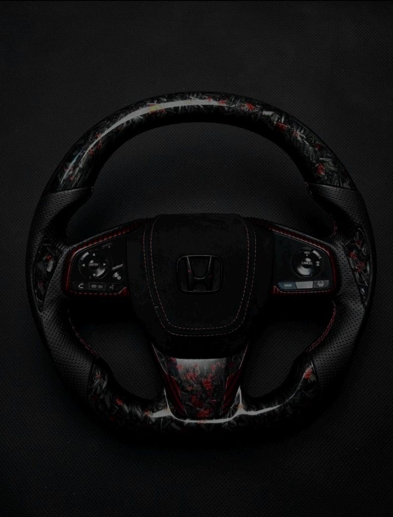 Puede incluir: Un volante Honda negro y rojo con un dise&ntilde;o de fibra de carbono. El volante presenta un agarre de cuero perforado con costuras rojas y controles integrados. El centro del volante tiene un acabado de gamuza negra con el logotipo de Honda.