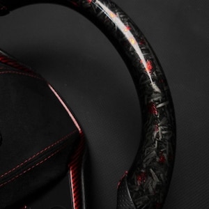 Puede incluir: Primer plano de un volante de fibra de carbono negro y rojo. El volante tiene un acabado brillante con detalles en rojo. El interior presenta cuero negro con costuras rojas y botones de control.