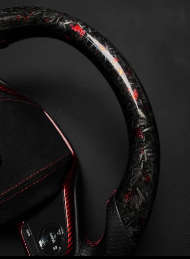 Puede incluir: Primer plano de un volante de fibra de carbono negro y rojo. El volante tiene un acabado brillante con detalles en rojo. El interior presenta cuero negro con costuras rojas y botones de control.