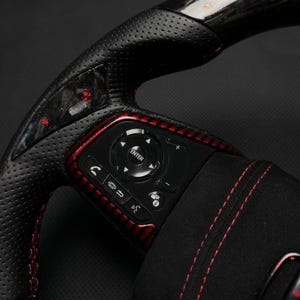 Puede incluir: Primer plano de un volante de coche negro y rojo. El volante presenta cuero perforado, detalles de fibra de carbono roja y botones de control. La consola central tiene un bot&oacute;n "ENTER" y otros botones de funci&oacute;n.