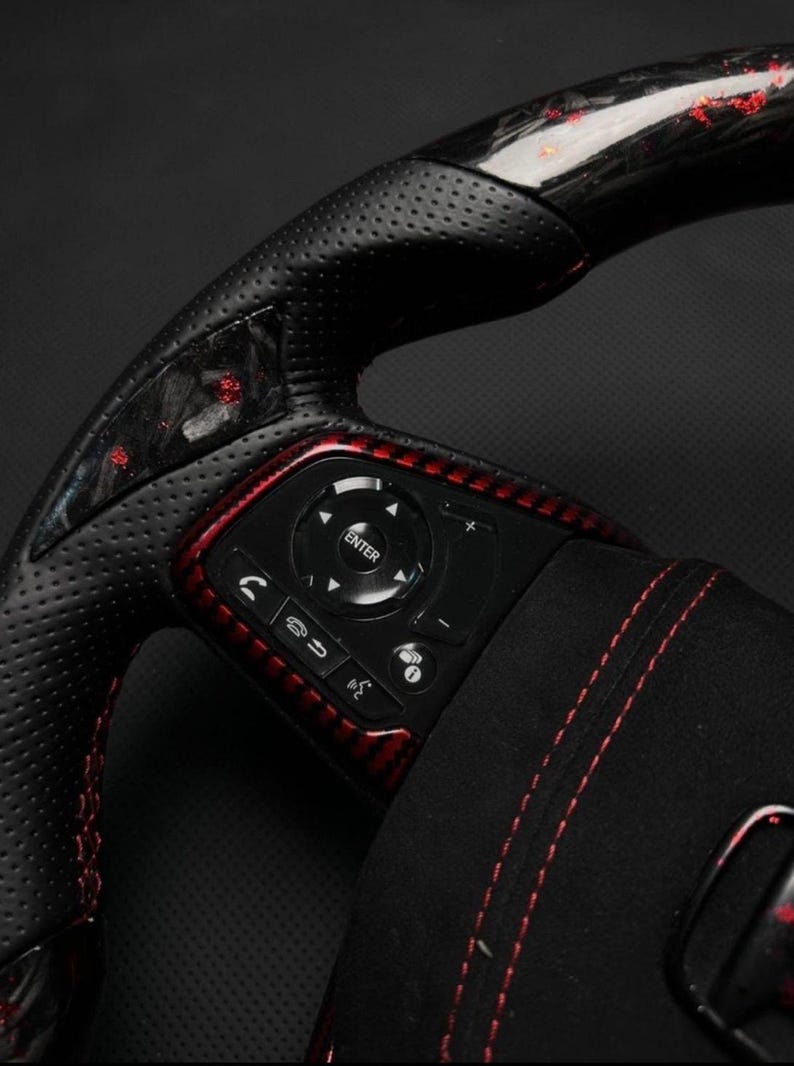 Puede incluir: Primer plano de un volante de coche negro y rojo. El volante presenta cuero perforado, detalles de fibra de carbono roja y botones de control. La consola central tiene un bot&oacute;n "ENTER" y otros botones de funci&oacute;n.