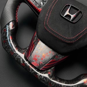Puede incluir: Primer plano de un volante de coche personalizado. El volante presenta un agarre de cuero negro perforado con costuras rojas, detalles de fibra de carbono y un centro de gamuza negra con el logotipo de Honda. El volante tiene controles integrados.