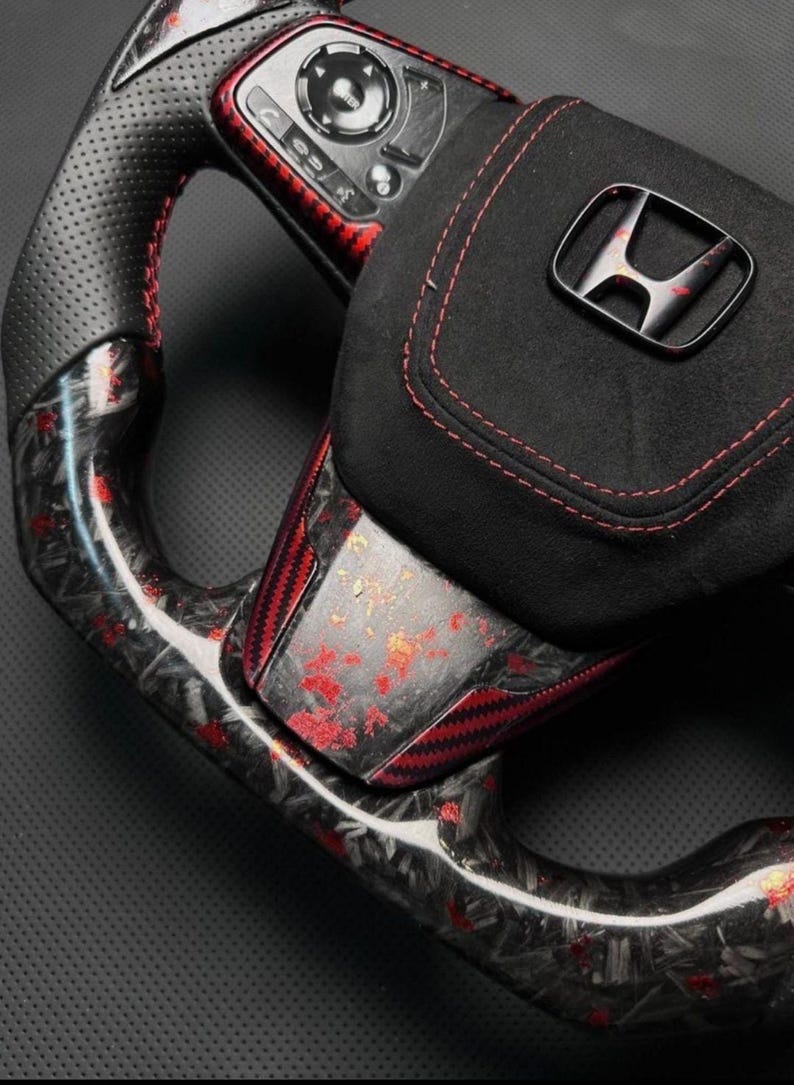 Puede incluir: Primer plano de un volante de coche personalizado. El volante presenta un agarre de cuero negro perforado con costuras rojas, detalles de fibra de carbono y un centro de gamuza negra con el logotipo de Honda. El volante tiene controles integrados.