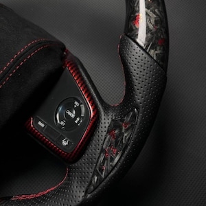 Puede incluir: Primer plano de un volante negro y rojo. El volante presenta un agarre de cuero perforado con costuras rojas. La secci&oacute;n superior tiene un dise&ntilde;o de fibra de carbono con detalles en rojo. Un panel de control con botones es visible.