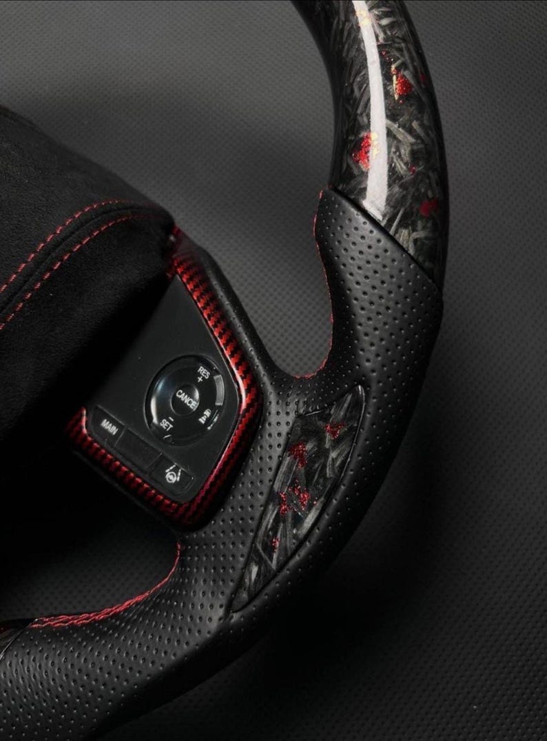 Puede incluir: Primer plano de un volante negro y rojo. El volante presenta un agarre de cuero perforado con costuras rojas. La secci&oacute;n superior tiene un dise&ntilde;o de fibra de carbono con detalles en rojo. Un panel de control con botones es visible.