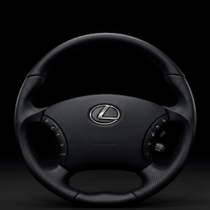 Puede incluir: Un volante de cuero negro con un agarre perforado y controles integrados. El centro presenta un emblema plateado de Lexus. El volante está sobre un fondo oscuro, resaltando su diseño y características.
