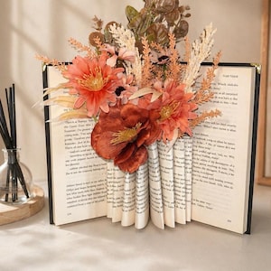 May include: An open book with pages fanned out to form a vase shape, filled with orange and peach-colored artificial flowers and foliage. The book has a black cover and gold-colored edges, creating a unique decorative piece.