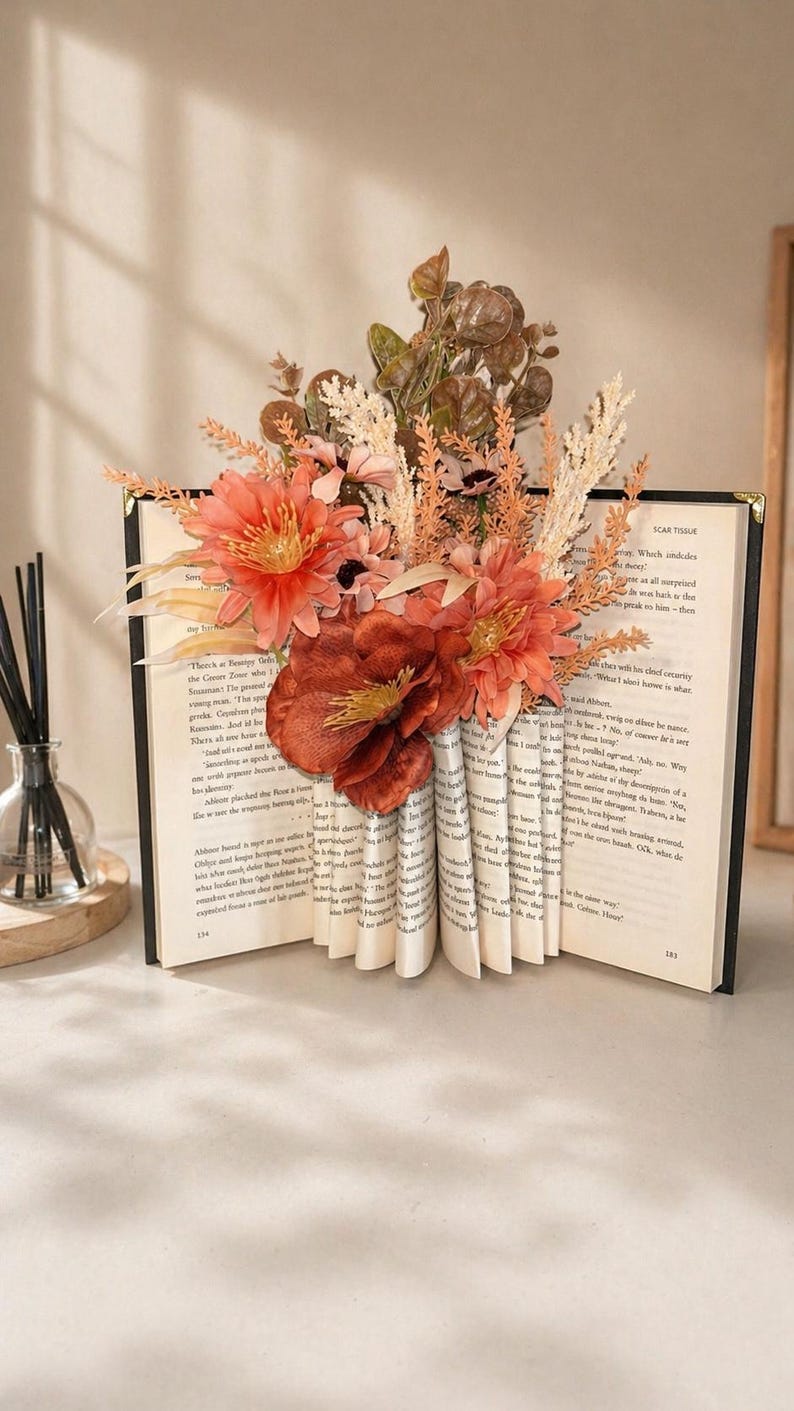May include: An open book with pages fanned out to form a vase shape, filled with orange and peach-colored artificial flowers and foliage. The book has a black cover and gold-colored edges, creating a unique decorative piece.