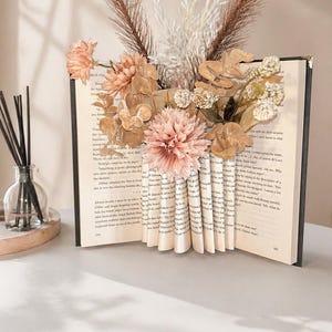 May include: A decorative book with folded pages forming a vase, filled with dried flowers and foliage in shades of peach, brown, and white. The book is open, revealing printed text, and sits on a white surface. A diffuser is in the background.