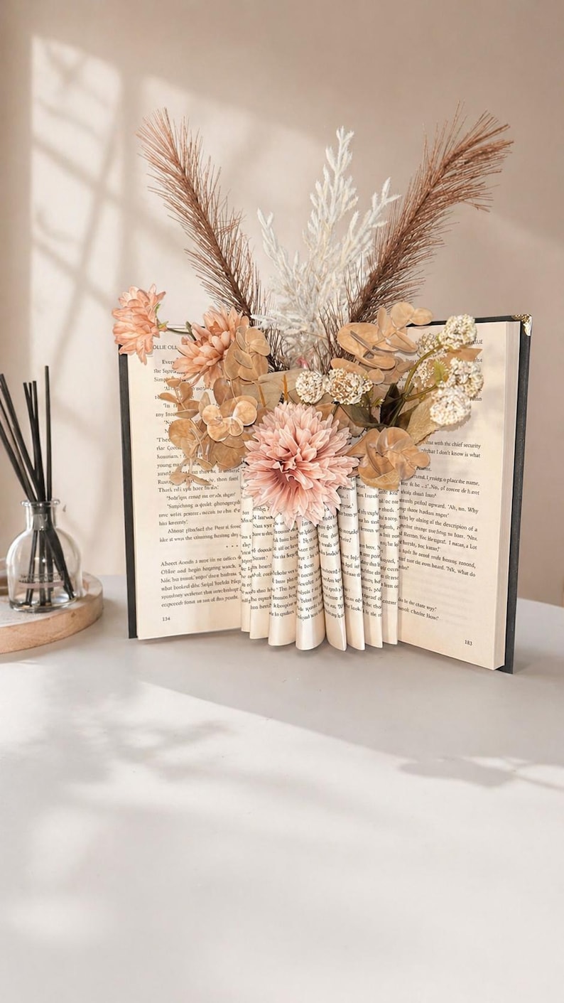 May include: A decorative book with folded pages forming a vase, filled with dried flowers and foliage in shades of peach, brown, and white. The book is open, revealing printed text, and sits on a white surface. A diffuser is in the background.