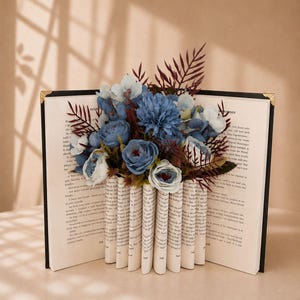 May include: A decorative book with pages folded to create a vase shape, holding a bouquet of blue and white flowers. The book has a black cover with gold corner accents. The background is a neutral beige.
