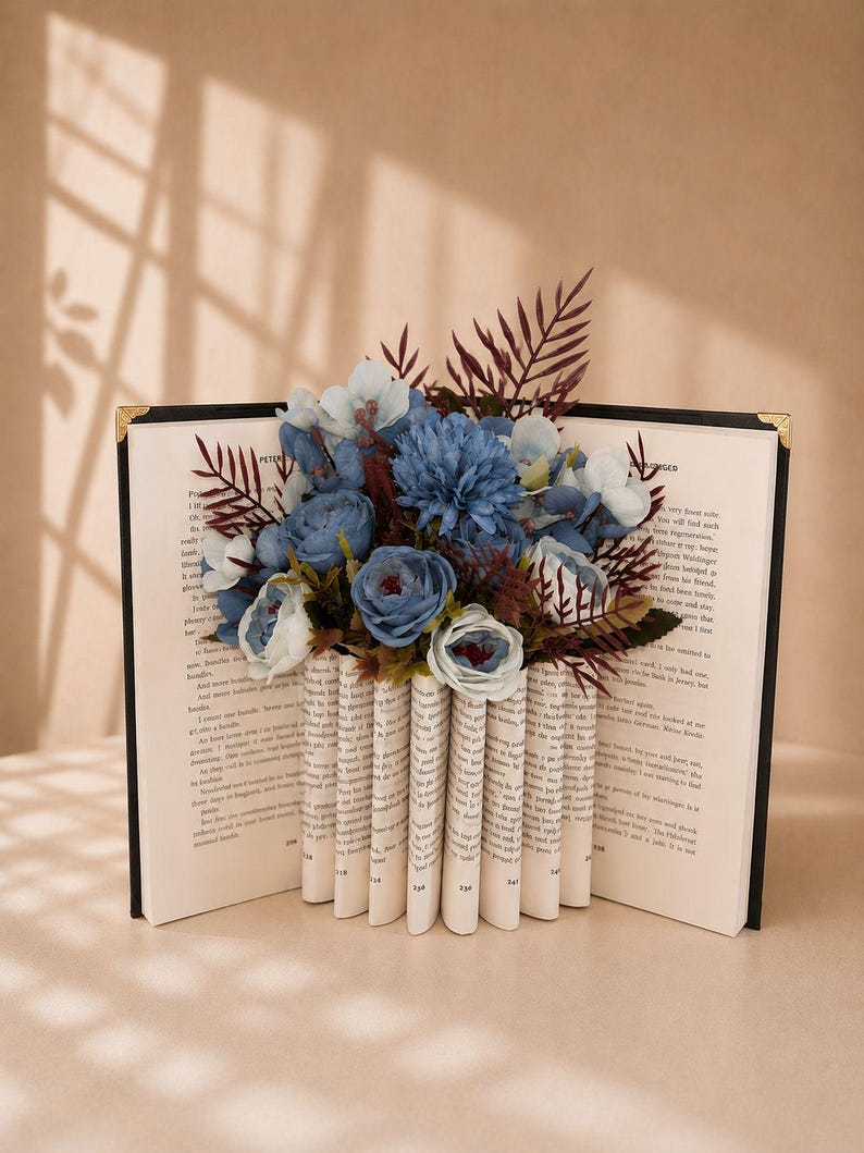 May include: A decorative book with pages folded to create a vase shape, holding a bouquet of blue and white flowers. The book has a black cover with gold corner accents. The background is a neutral beige.