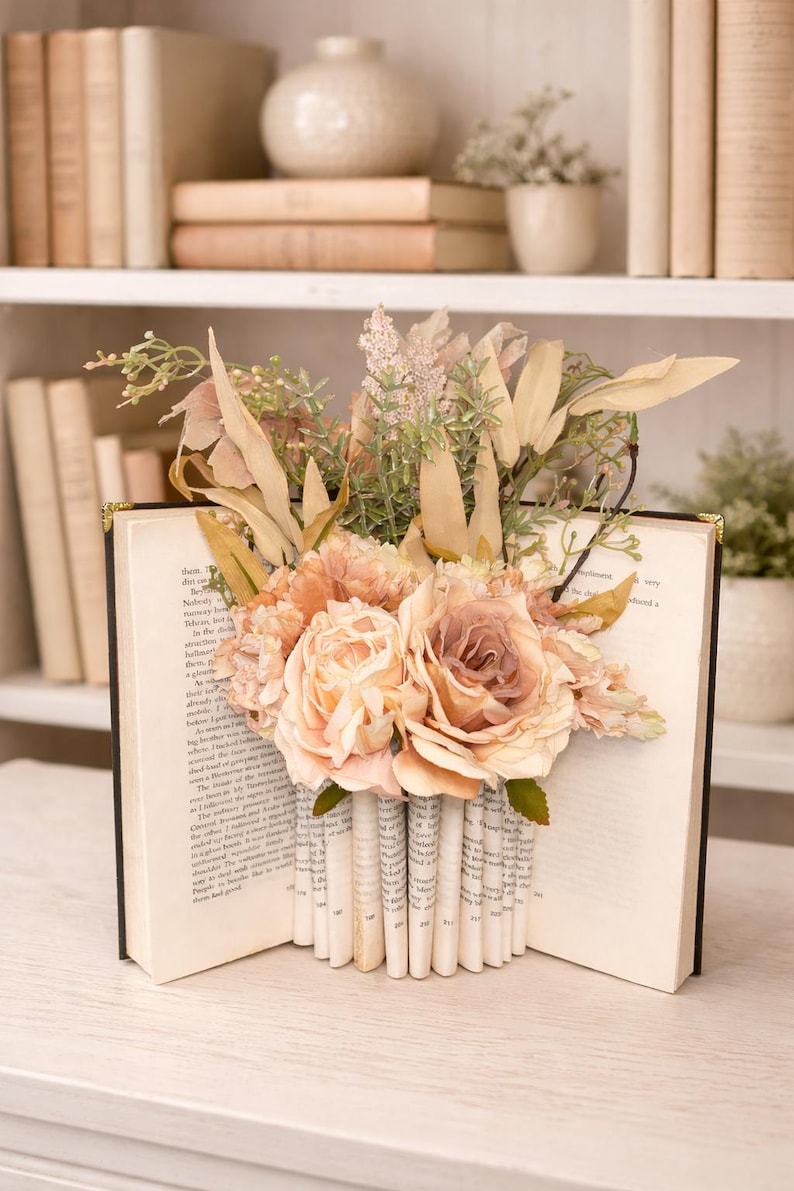 May include: An open book with pages folded to create a vase shape, filled with peach and cream-colored roses and greenery. The book is set against a backdrop of a white bookshelf with other books and decorative items.