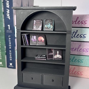 Könnte beinhalten: Ein schwarzes Miniatur-Bücherregal mit gewölbtem Oberteil und zwei unteren Schubladen. Die Regale enthalten winzige Buchnachbildungen, darunter Six of Crows, Siege and Storm, A Feast for Crows, A Storm of Swords, The Night Circus, Twilight und Breaking Dawn.