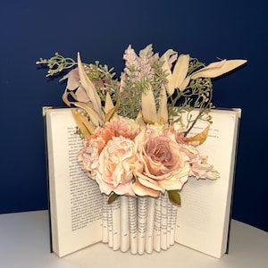 May include: A decorative book with pages folded to create a vase shape, filled with peach and cream-colored roses and other faux flowers. The book is open, revealing printed text, and sits on a white surface against a dark blue background.