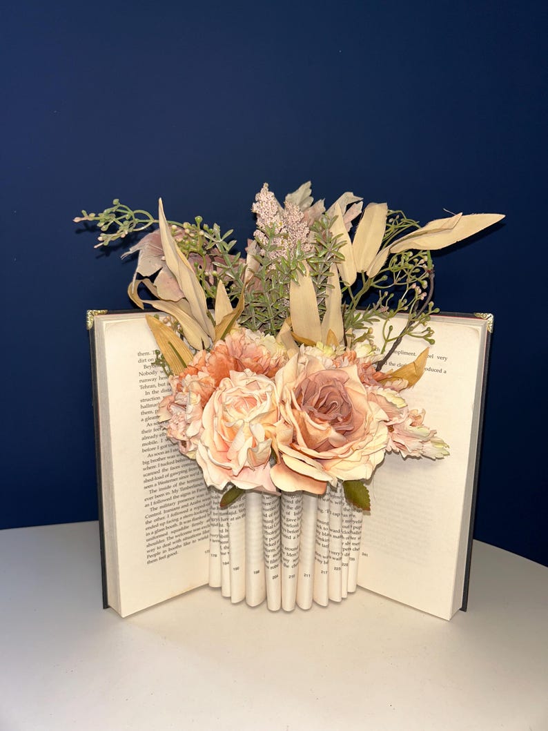 May include: A decorative book with pages folded to create a vase shape, filled with peach and cream-colored roses and other faux flowers. The book is open, revealing printed text, and sits on a white surface against a dark blue background.