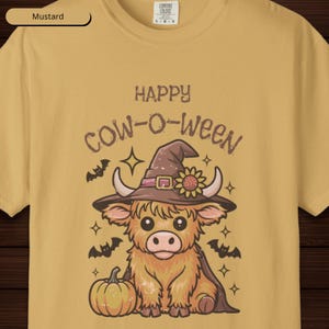 May include: Mustard-coloured t-shirt with a Halloween graphic. The design features a cartoon cow wearing a witch hat and cape, with the text "HAPPY COW-O-WEEN". A pumpkin, bats, and stars complete the festive design.