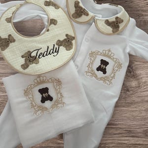 Peut inclure: Un ensemble de vêtements de bébé blanc comprenant un bavoir, une combinaison et une couverture. Le bavoir a une bordure beige et est décoré d'ours en peluche bruns et du nom "Teddy". La combinaison et la couverture ont un motif d'écusson d'ours en peluche brun.