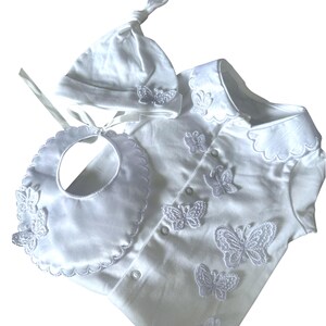 Peut inclure: Ensemble de vêtements de bébé blanc comprenant une chemise boutonnée, un bavoir et un bonnet noué. L'ensemble est orné d'applications de papillons blancs. Le bavoir a un bord festonné.
