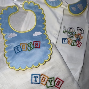 Puede incluir: Un body blanco para bebé con cuello azul bordado con nubes, un babero festoneado azul claro con nubes y letras en bloque bordadas que forman TOYS, y un paño de muselina blanco con letras en bloque bordadas que forman TOYS.