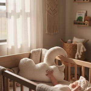 May include: A cream-coloured, plush swan-shaped pillow sits in a wooden cot. The nursery has a neutral colour palette, with a macrame wall hanging and wooden shelves. A baby is lying in the cot, with a hand resting on the pillow.