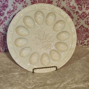 May include: A cream-colored ceramic egg serving platter featuring twelve oval indentations arranged in a circle. The center and the outer rim are decorated with embossed floral patterns. The platter is held upright by a thin, dark metal stand.